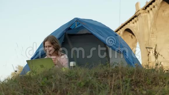 年轻女子在旧桥附近的旅游帐篷里的笔记本电脑后面工作视频的预览图
