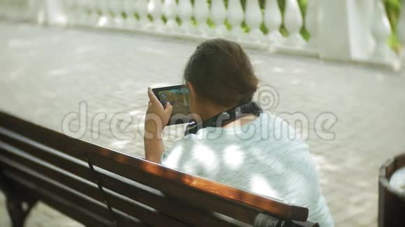 一个夏天一个小胖女孩坐在长凳上听音乐或看视频带着平板电脑和耳机视频的预览图