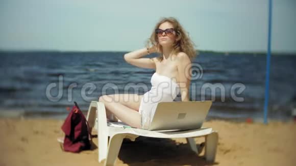 一个穿着白色比基尼的年轻女孩躺在沙滩上的躺椅上晒太阳正在使用笔记本电脑视频的预览图