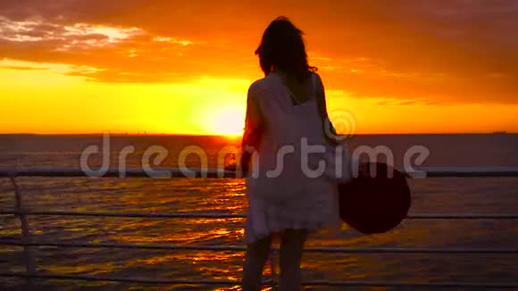 美丽的日落时分穿着白色连衣裙和戴着帽子的年轻迷人女子在海上背景下摆姿势视频的预览图