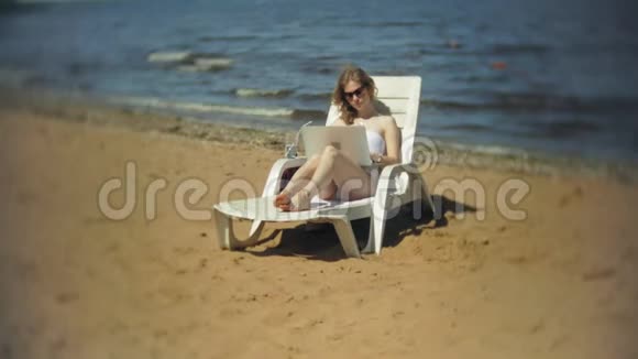 一个穿着白色比基尼的年轻女孩躺在沙滩上的躺椅上晒太阳正在使用笔记本电脑视频的预览图