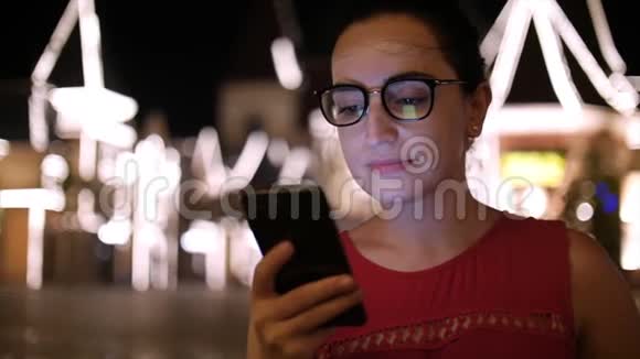 在夜市里在背景夜市里一个迷人的女孩一边用手机在街上走来走去视频的预览图
