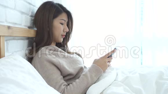 快乐的亚洲女人早上在床上使用智能手机视频的预览图