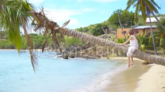可爱的小女孩坐在棕榈树在暑假白色海滩视频的预览图
