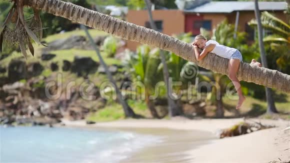 可爱的小女孩坐在棕榈树在暑假白色海滩视频的预览图