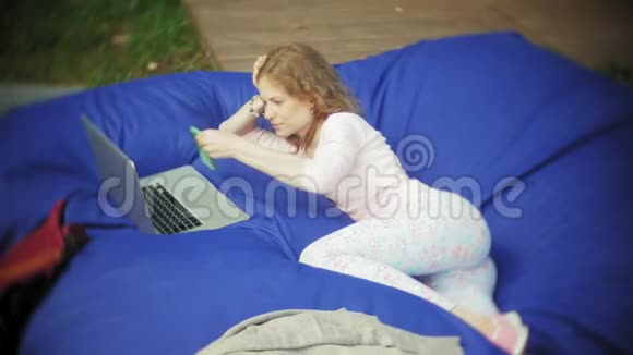 年轻女子用手提电脑躺在公园的一个休息区软泡芙上视频的预览图