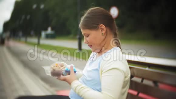 靠近可爱的小胖女孩双手坐在公园的长凳上吃蛋糕视频的预览图