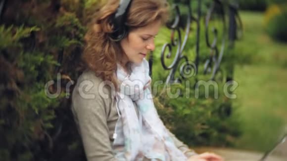 有笔记本电脑的女人在一个美丽的绿色公园的长凳上放松一个常年在树洞里工作的年轻女人视频的预览图