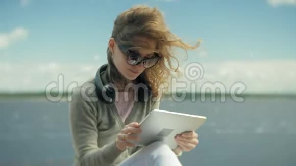 年轻的美女戴着耳机坐在沙滩上用平板电脑视频的预览图