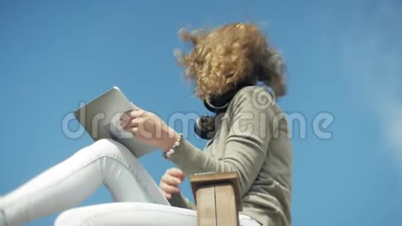 年轻的美女戴着耳机坐在沙滩上用平板电脑视频的预览图