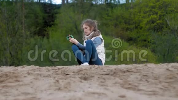 美丽的女孩微笑着用智能手机在河岸上自拍视频的预览图