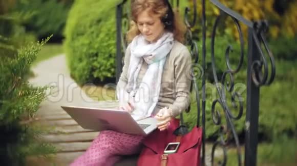 有笔记本电脑的女人在一个美丽的绿色公园的长凳上放松一个常年在树洞里工作的年轻女人视频的预览图