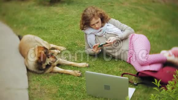 一个拿着笔记本电脑的女人和她的狗在一个美丽的绿色公园的草地上放松一个常年年轻的女人视频的预览图