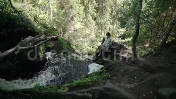 年轻女子坐在瀑布附近的岩石上视频的预览图