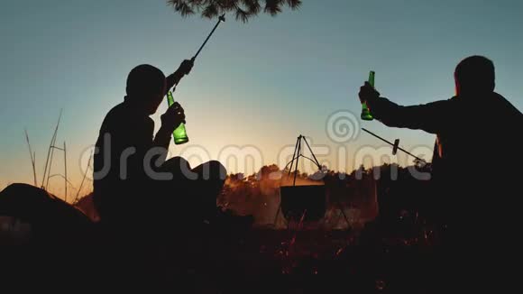 两名游客正坐在篝火晚会上在大自然中露营剪影阳光日落两个男人在度假时剪影视频的预览图