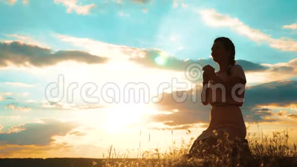 女孩在日落时双手交叉在生活方式祈祷剪影中女人跪着祈祷慢动作视频女孩视频的预览图