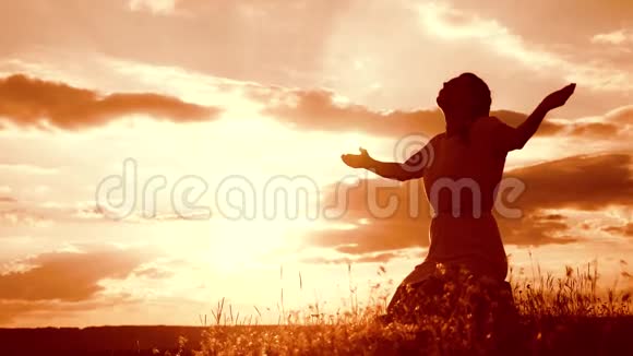 女孩在日落时双手交叉祈祷剪影女人跪着祈祷慢动作视频概念视频的预览图