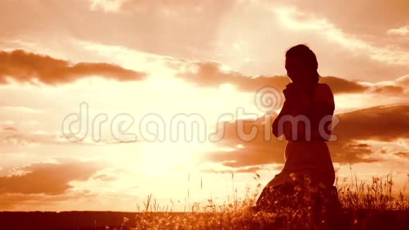 女孩在日落时双手交叉祈祷剪影女人跪着祈祷慢动作视频概念视频的预览图