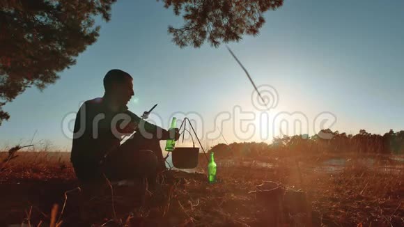 男游客坐在篝火旁喝啤酒在大自然野营的剪影下晒日落度假男士剪影视频的预览图