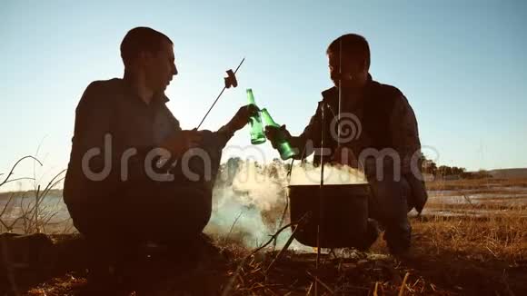 两位游客啤酒是坐在篝火旁的生活方式在大自然野营的剪影阳光下日落两个人视频的预览图