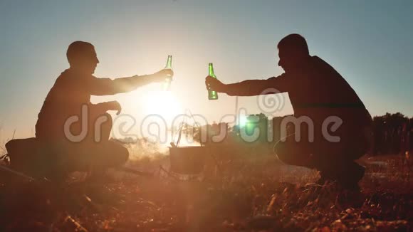 两位游客啤酒是坐在篝火旁的生活方式在大自然野营的剪影阳光下日落两个人视频的预览图