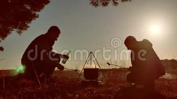 两位游客啤酒正坐在篝火徒步聚会上在大自然中露营剪影阳光夕阳的生活方式两个人视频的预览图