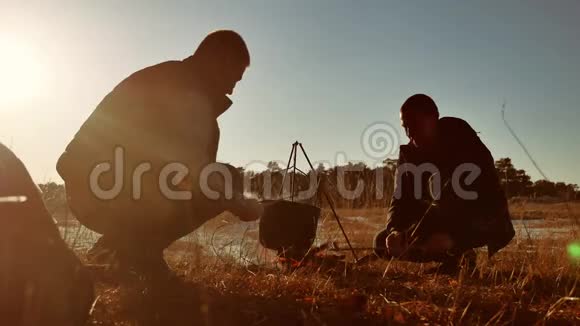 两位游客啤酒是生活方式坐在篝火徒步聚会上在大自然中露营剪影阳光日落两个人视频的预览图