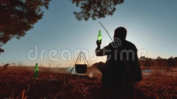 男子游客是啤酒坐在篝火晚会上在大自然中露营剪影阳光日落户外男子剪影视频的预览图