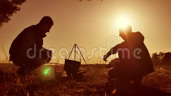 两位游客啤酒正坐在篝火生活方式的帽子派对上在大自然中露营剪影阳光日落两个人视频的预览图