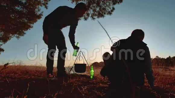 男游客坐在篝火旁喝啤酒在大自然野营的剪影下晒日落度假男士剪影视频的预览图