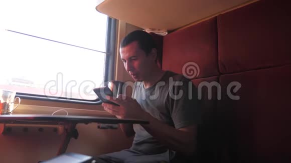 一名男子坐在火车车厢里手里拿着一部智能手机喝着咖啡和茶生活方式慢动作视频的预览图