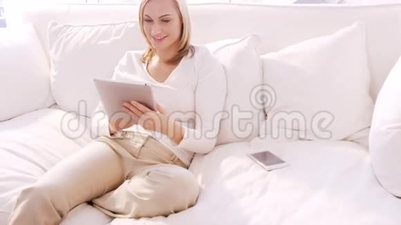 金发女人微笑着在沙发上用平板电脑视频的预览图
