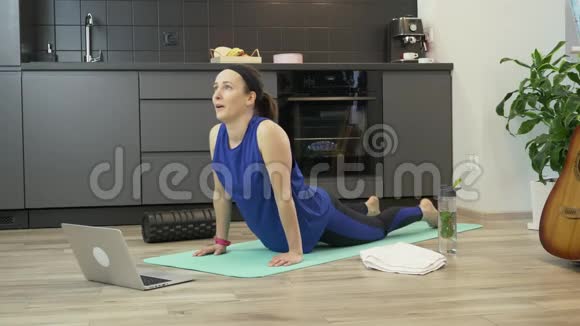 年轻女孩在厨房里做伸展运动和瑜伽练习使用笔记本电脑上的在线健身应用程序女子体育用品视频的预览图