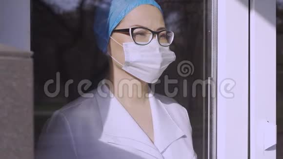 呼吸保健和生命安全covid19病毒女医生戴着外科口罩穿着制服透过窗户视频的预览图