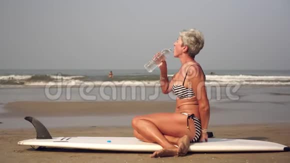 一个身材好的成熟女人坐在沙滩上拿着冲浪板和喝水视频的预览图