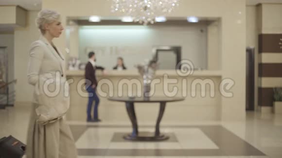 自信的白人金发女人带着手提箱经过酒店的前厅有模糊的人在前台交谈视频的预览图