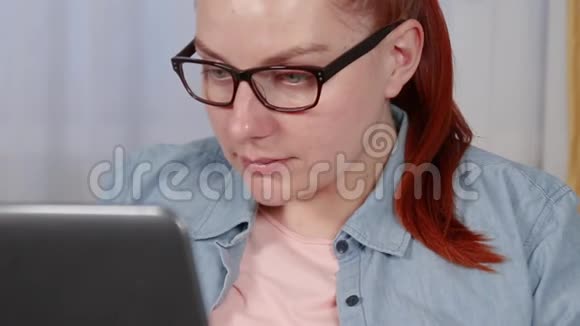漂亮的年轻女子戴着眼镜穿着一件衬衫坐在客厅里的笔记本电脑上工作视频的预览图