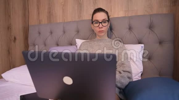 一幅漂亮的自由职业者女孩戴着眼镜的画像床上有一台笔记本电脑在一里后面有透明的墙视频的预览图