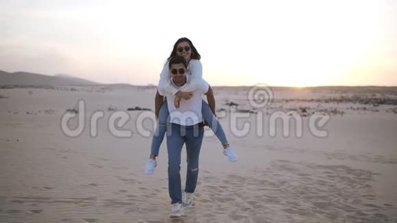 快乐可爱的一对成年夫妇穿着白色衬衫和牛仔裤男人背着女朋友在沙漠中奔跑视频的预览图
