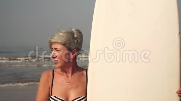 成熟的女人在海滩上拿着冲浪板祖母在大海的背景上冲浪视频的预览图