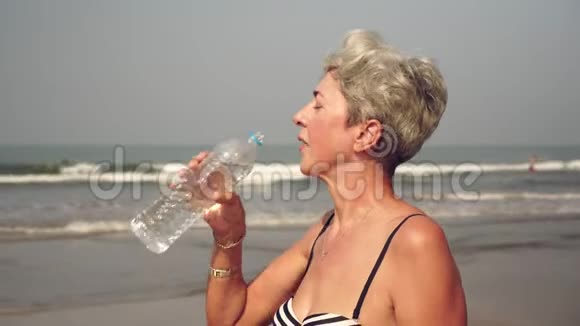 成熟迷人的女人从海滩上的瓶子里喝水视频的预览图