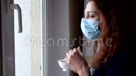 年轻女子戴着医疗面具喝咖啡在隔离期间看着窗外社会距离视频的预览图