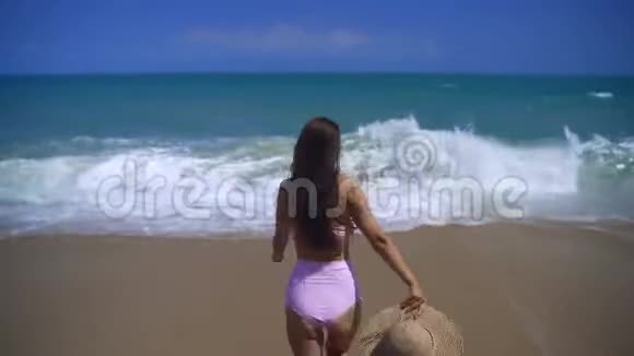 一个穿着粉红色泳衣的漂亮女孩脱下帽子朝海浪跑去视频的预览图