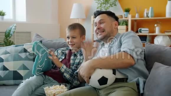 足球迷爸爸和孩子在家看电视比赛手里拿着泡沫塑料球视频的预览图