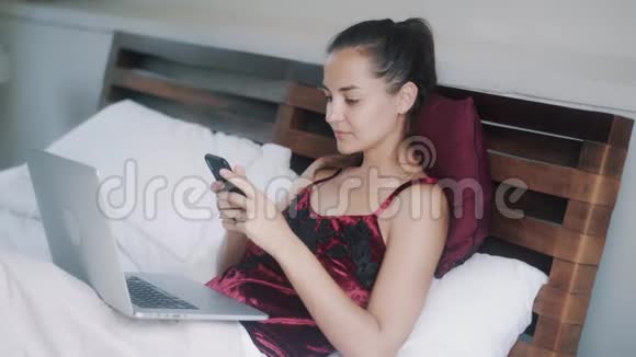 妇女躺在床上在大流行病期间使用电话和笔记本电脑在家工作视频的预览图