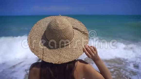 一个戴草帽的女孩看着海浪女孩用手拿着帽子从后面的特写镜头视频的预览图