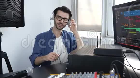 录音室的年轻人坐在桌子旁调整混合控制台通过耳机听音乐视频的预览图