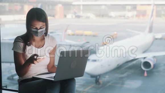 戴防护医疗面罩的妇女在机场使用笔记本电脑和智能手机视频的预览图