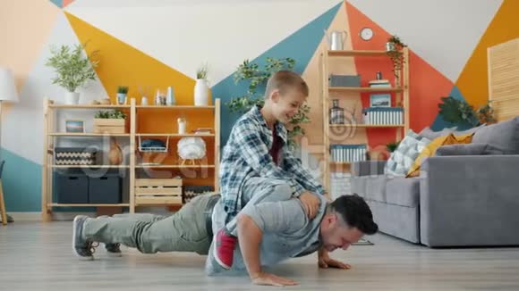 强壮的男人父亲做俯卧撑而儿子则坐在他的背上在家里微笑视频的预览图