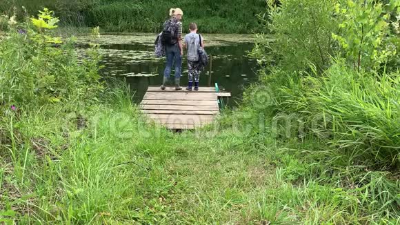 母亲和儿子带着背包的游客徒步旅行者沿着长满青草的小路走到河边欣赏大自然视频的预览图
